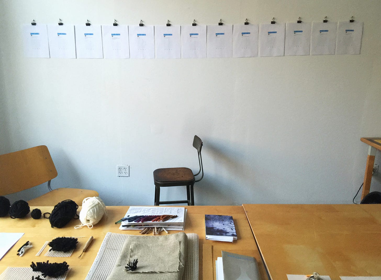 View of a studio space with a pair of wooden tables pushed together. On the tables are an array of materials including yarn, yarn samples, and open-weave canvas and books. Pinned up on the wall are a line of paper sheets with blue bars. You can't make them out in the photograph, but they are printed-out tweets.