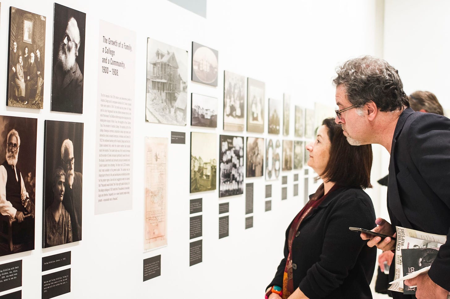 Two gallery visitors are shown examining photographs and historical documents in a white gallery space.