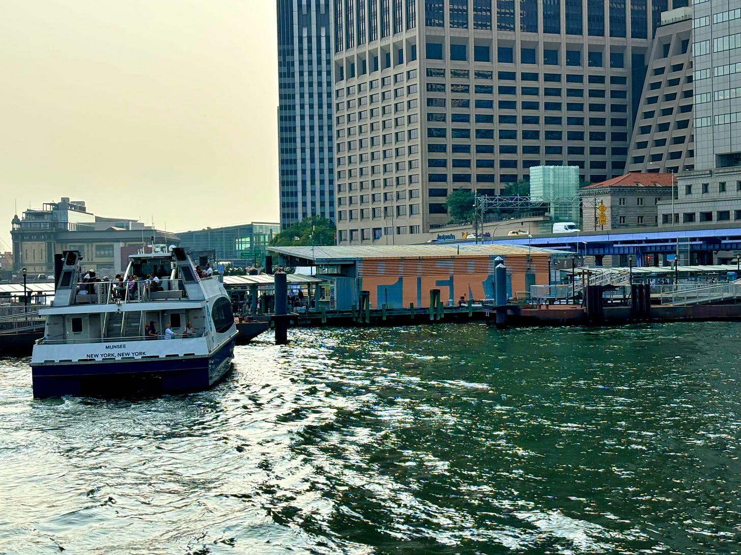 An NYC Ferry pulls into Pier 11. Tall Wall street buildings are ranged behind the dock.