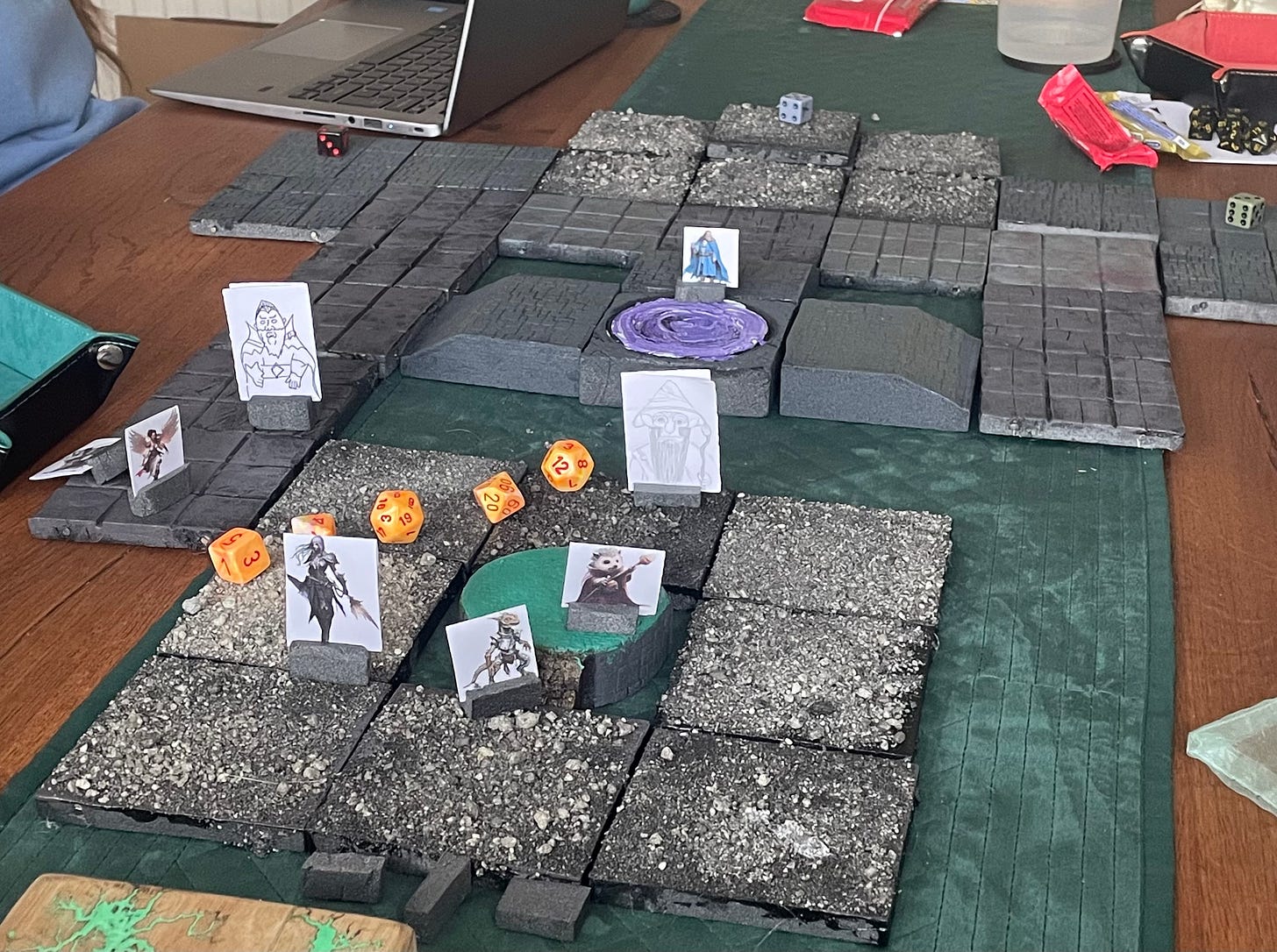 A number of stone tiles placed on a wooden table, to create a battleground. Several characters stand on a portal in the foreground, cut off from the portal in the middle of the room by a wall of fire represented by orange dice.