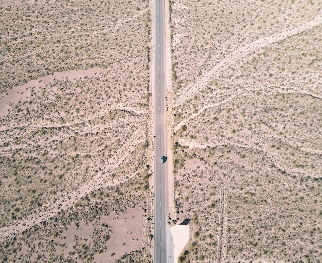 A desert with shrubbery. A highway runs vertically down the middle, and a lone car drives down it.