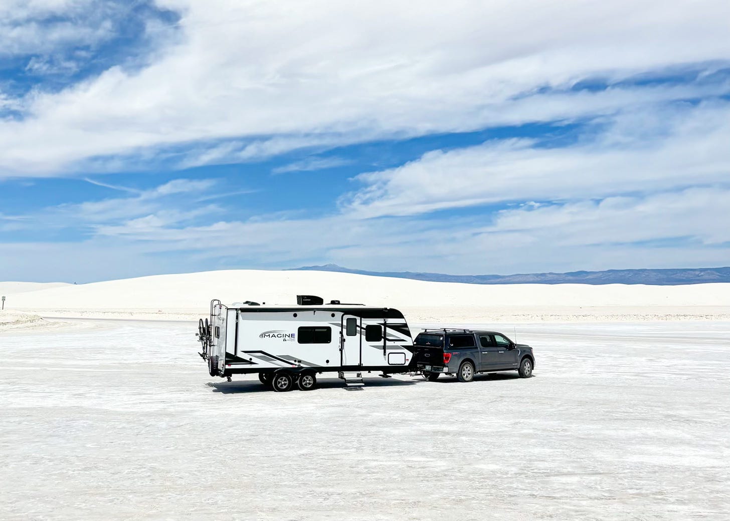 Grey truck pulling white and black travel trailer on wide expanse of white sand with bright blue sky and white clouds