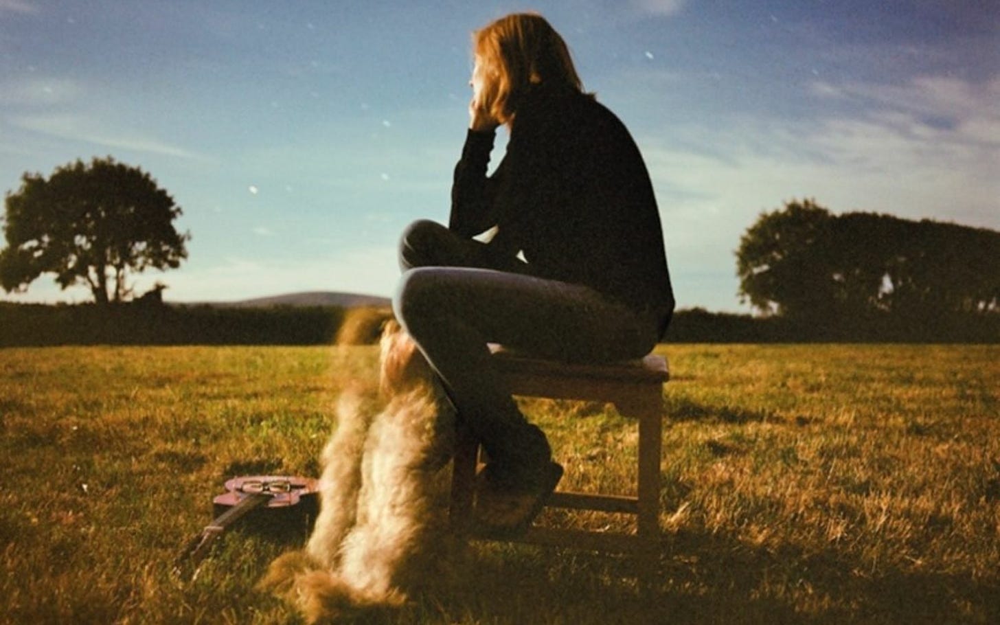 Mulher sentada num banco sem encosto, com um cachorro encostado nas pernas dela, um violão no gramado. A foto está desfocada, só tem natureza em volta e a artista olha para traz contemplando o céu azul