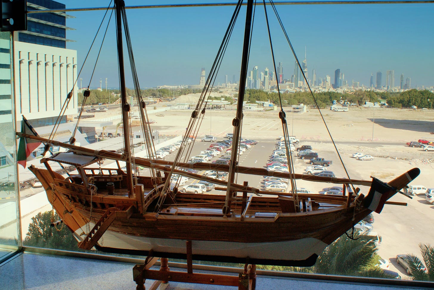 A large wooden model replica of an Arabian dhow sits by the window of a modern building overlooking a middle-eastern city skyline.