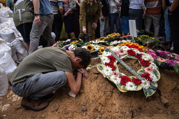 A man collapses on a dirt grave, on which several floral wreaths are laid.