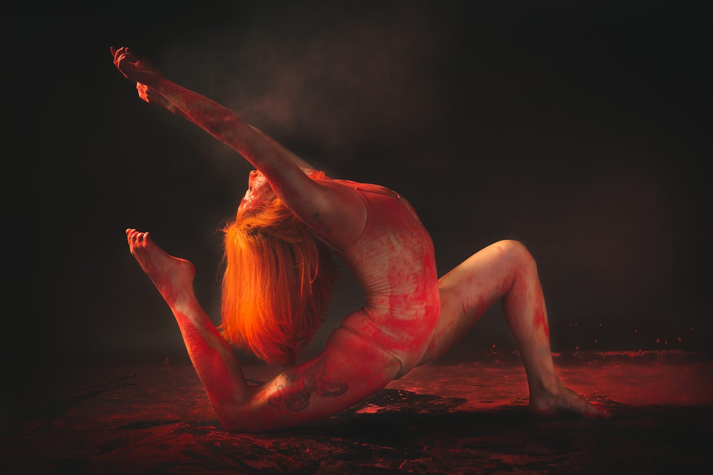 A red haired female acrobat covered in red pigment reached up to the sky from the earth.