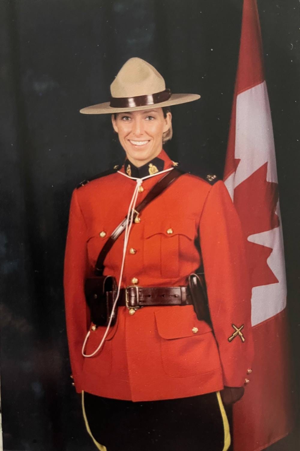 A woman in a red police uniform with a beige brimmed hat stands before a Canadian flag and smiles at the camera. 