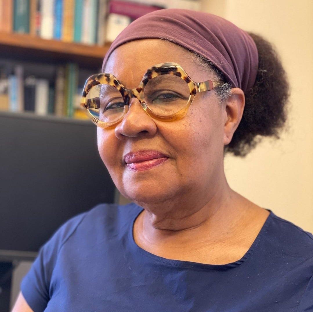 A black older woman in a light mauve headwrap, tortoise shell frame glasses and blue blouse sits in front of a book shelf (blurred in the background)
