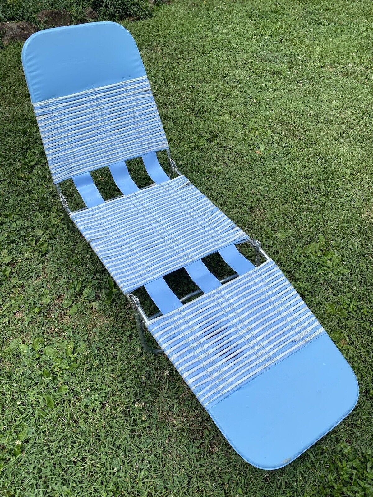 A folding lounge chair made of an aluminum tube frame and alternating blue and white plastic straps in a horizontal stripe pattern is shown outdoors on a green grass lawn.