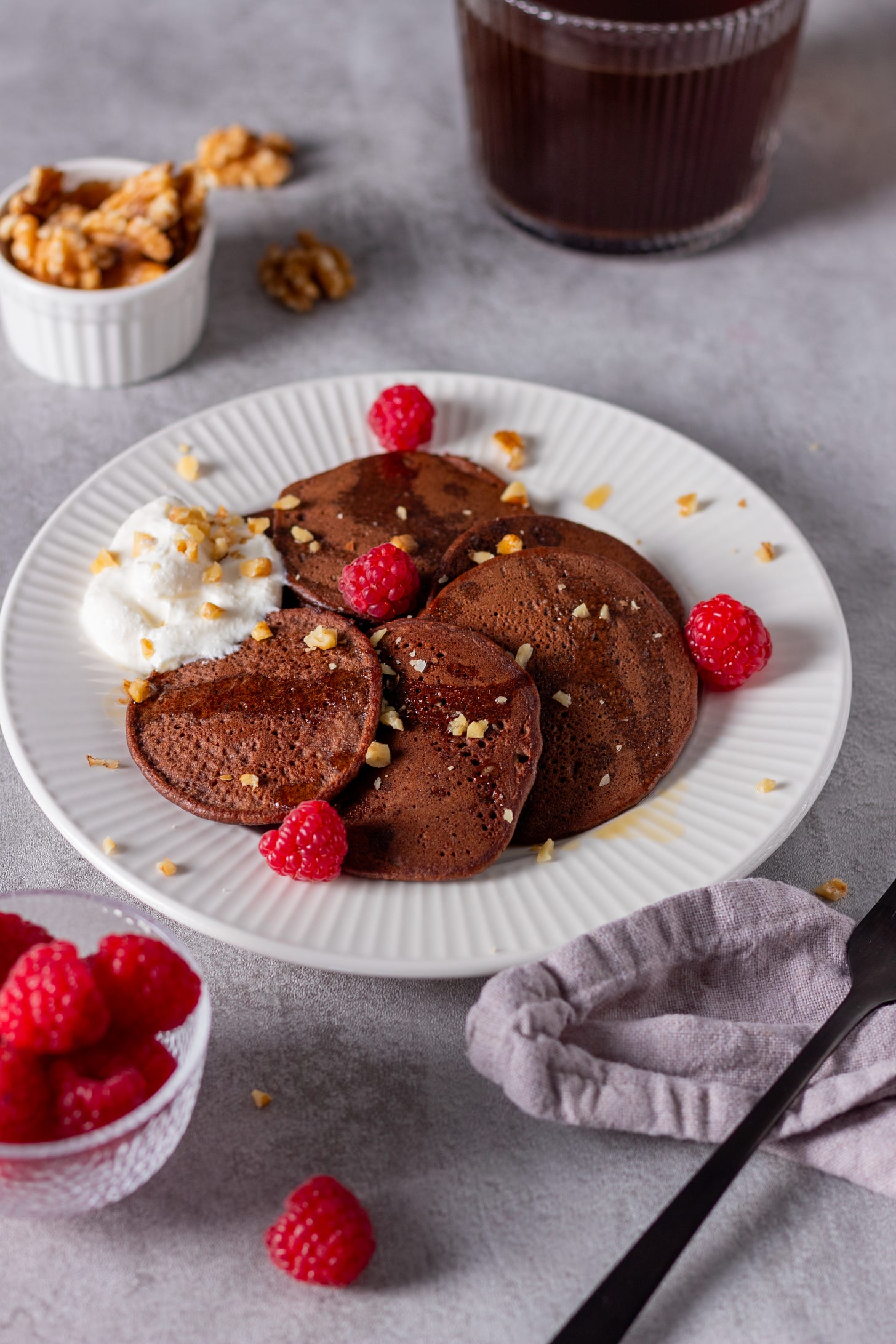 Plate of pancakes topped with syrup, chopped nuts, and fresh raspberries with a dollop of whipped cream on the side.