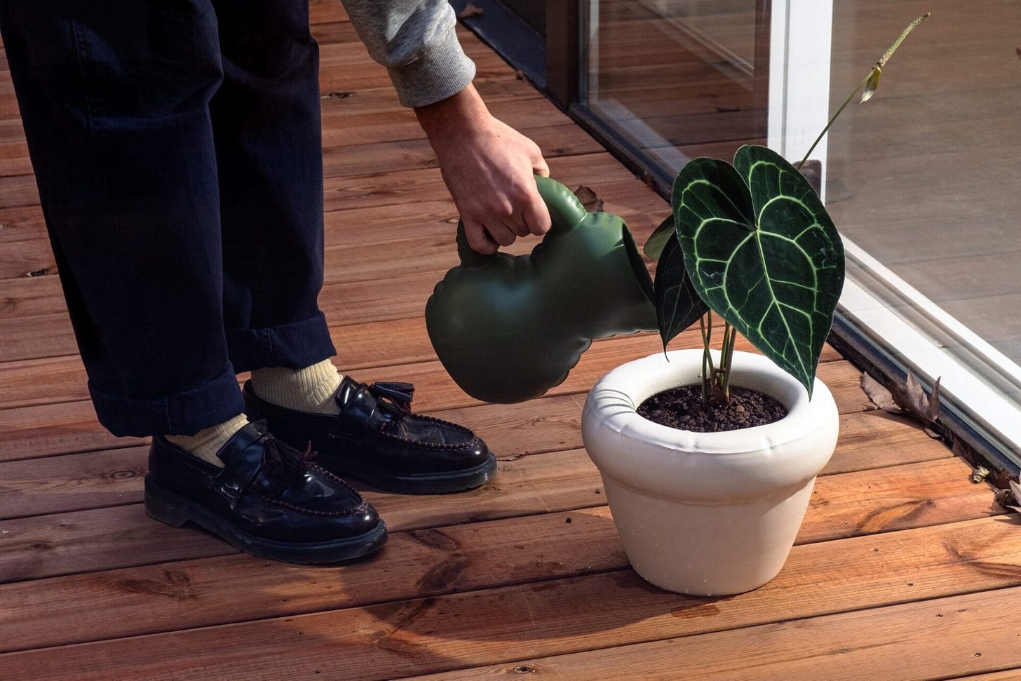 a person shown from the knees down pours water from a green pitcher into a plant in a white vase, both objects appearing inflated but actually made of ceramic