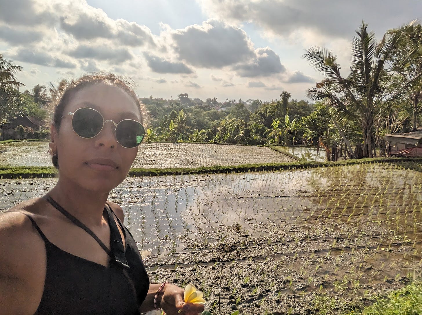 Nathalie poses in front of a rice field and countless trees. She is wearing round green tinted sunglasses and a black dresses and holds a yellow flower in her hand