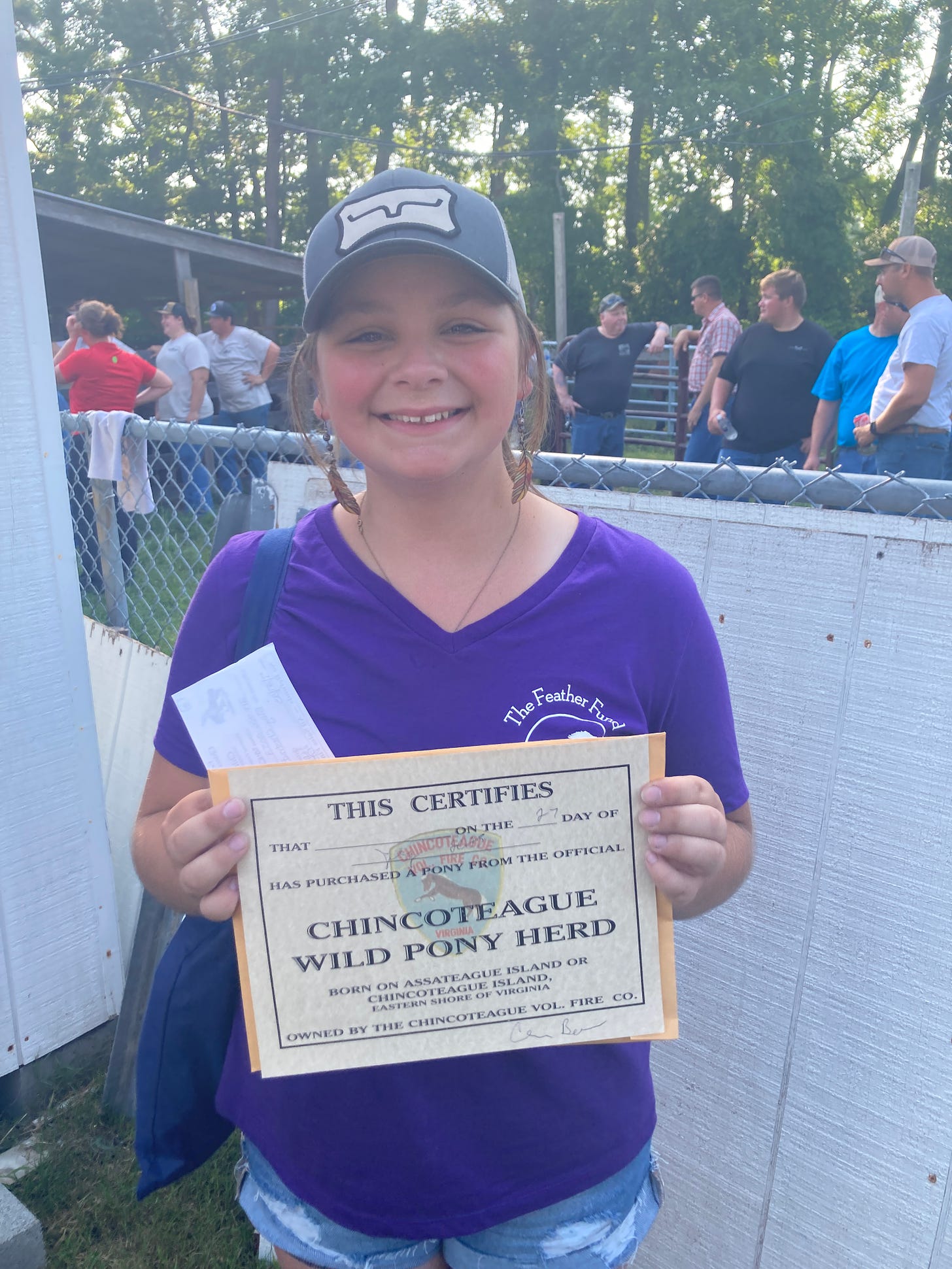  A teenage girl in a purple t shirt and blue hat, standing by a white fence, with a beige certificate that certifies her ownership of a member of the Chincoteage Wild Pony Herd