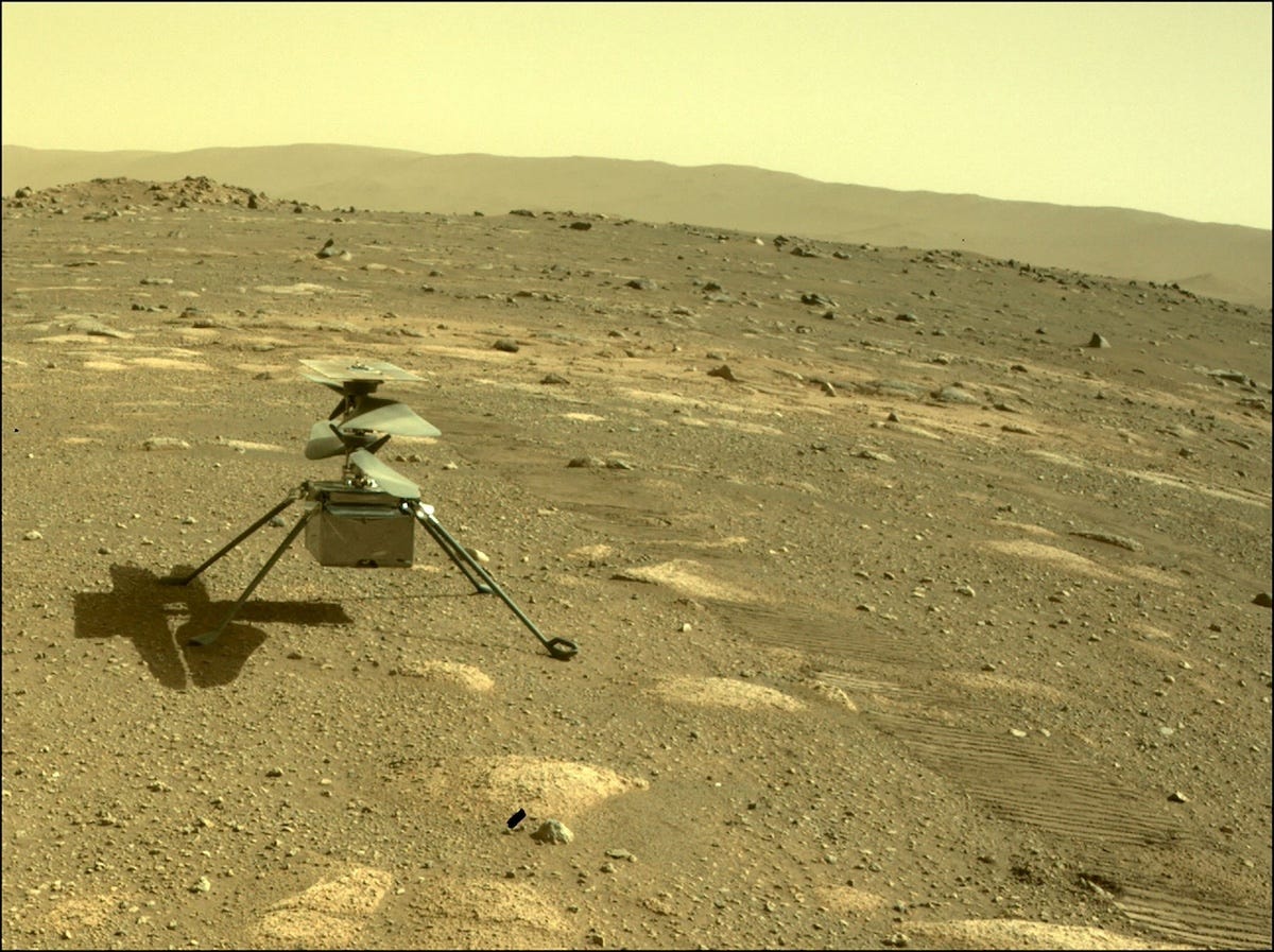 The image shows a small helicopter-like drone on a rocky, dusty landscape under a yellow-brown sky, resembling the surface of Mars.