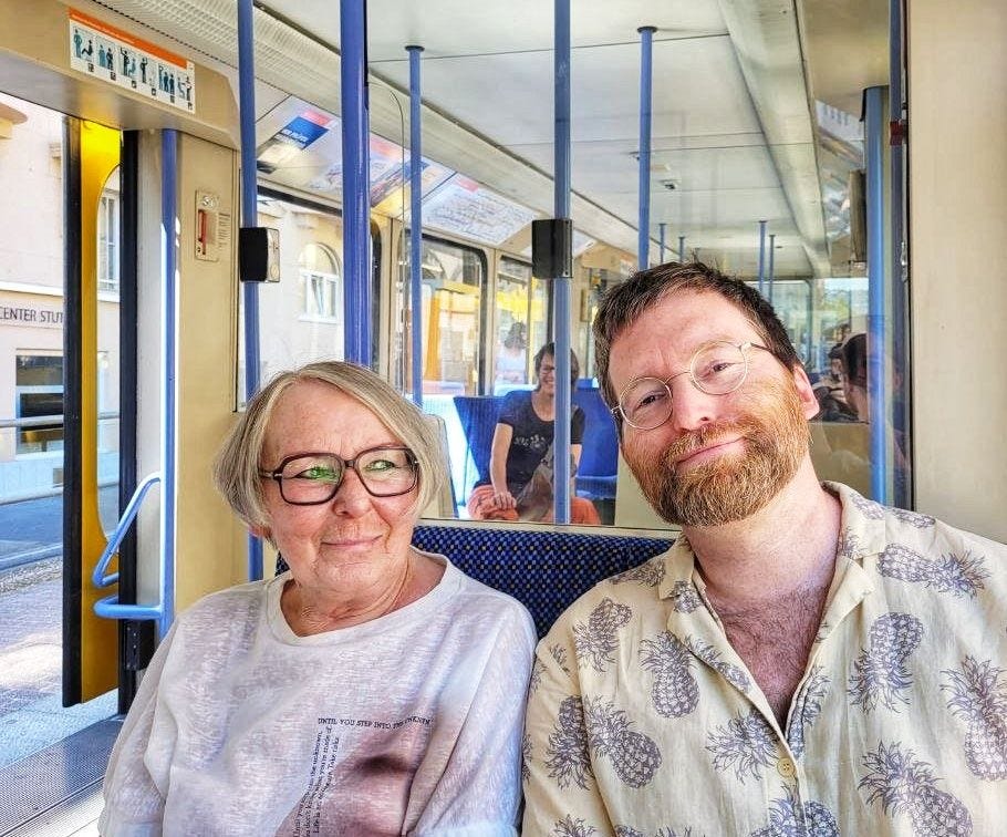 In sommerlicher Kleidung sitzen der Autor und seine Mutter nebeneinander in einer Stuttgarter Straßenbahn. Der Autor schaut in die Kamera, seine Mutter aus dem Fenster. Beide sind fröhlich.