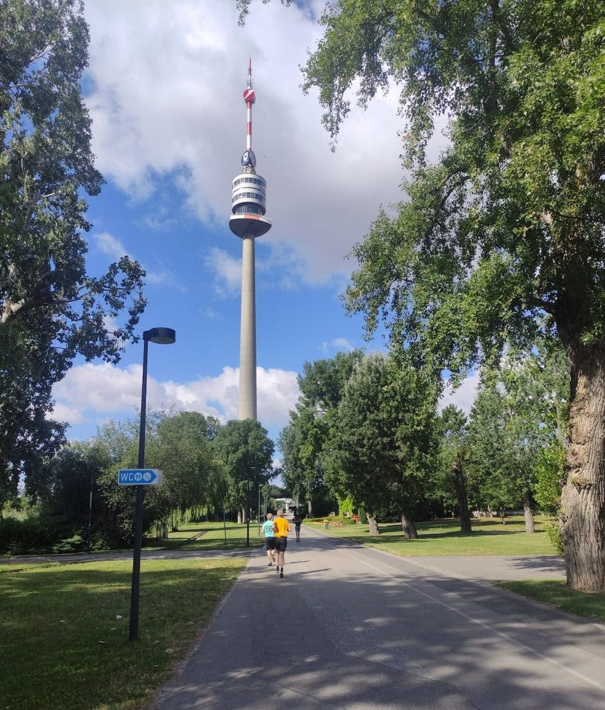 The tall Donauturm, or Danube tower, rises in the park and the path and runners head towards it.
