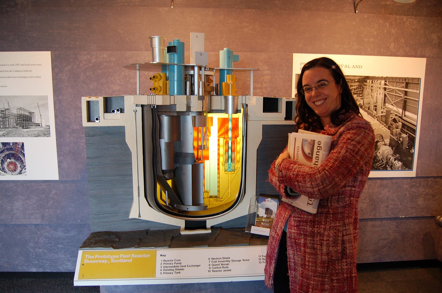 A dark haired woman in a bright pink coat standing next to a 3D cut out model of a nuclear reactor