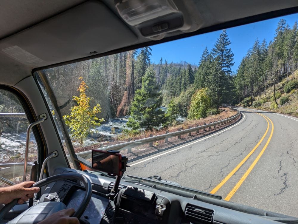 the view from inside Walter the truck, looking out at a narrow, winding highway alongside a whitewater river with blue sky and evergreen trees