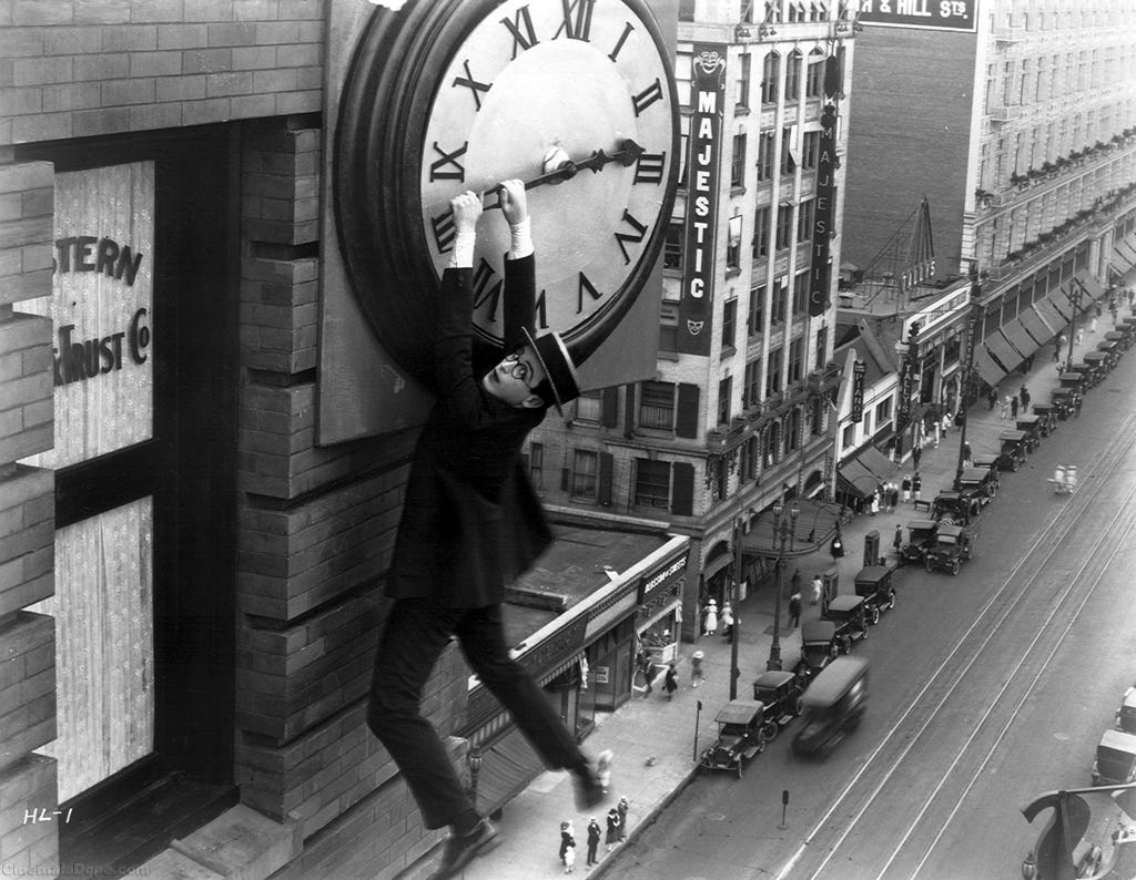 A black and white image of a silent movie. A man with specs and a flat, straw hat hangs off the hand of a clock on the side of a tall building in the city.