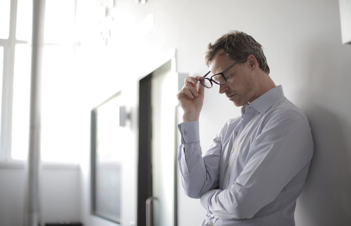 Free Thoughtful man in a bright room holding his glasses while leaning against a wall. Stock Photo