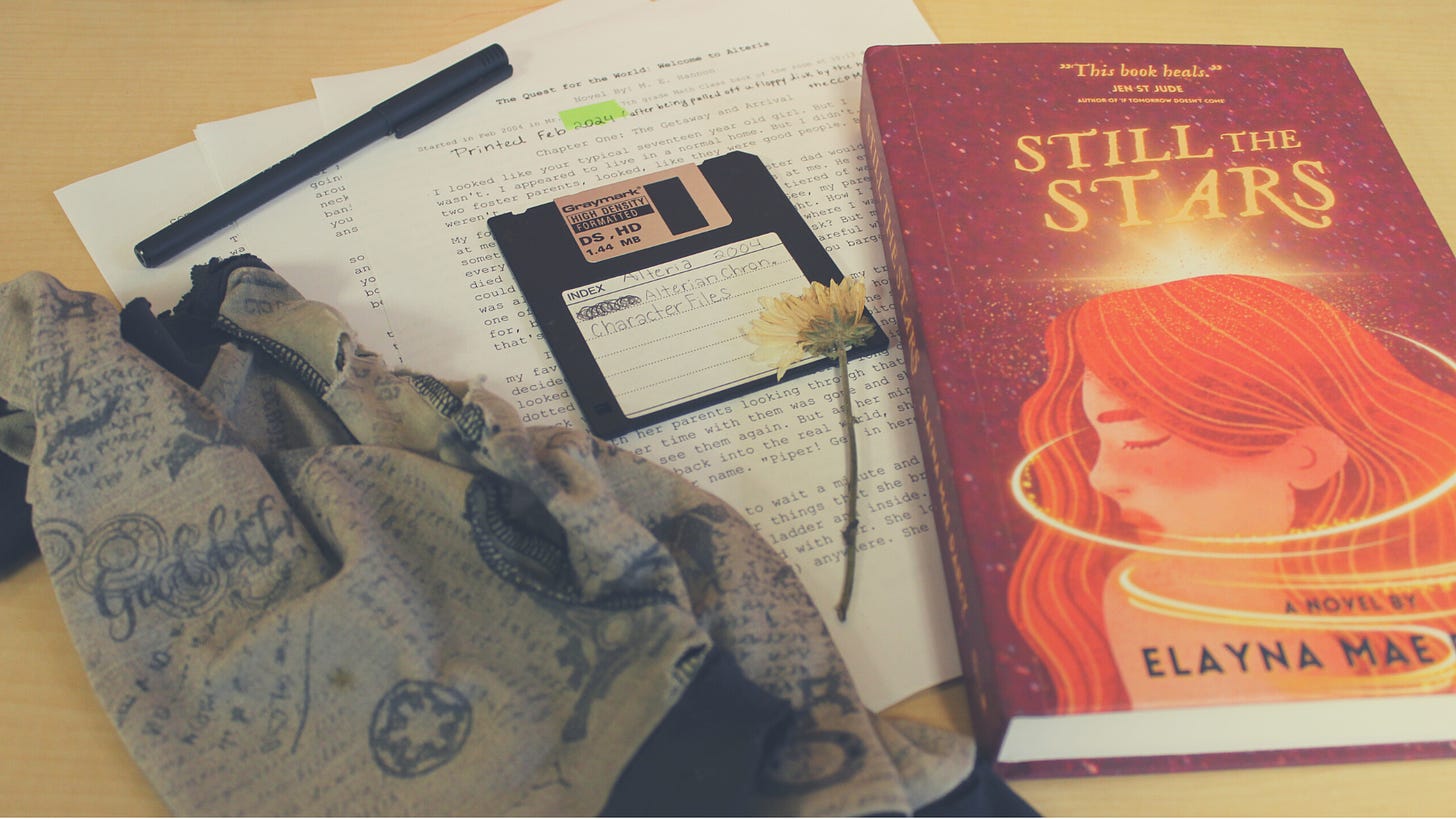 Photo of a copy of STILL THE STARS by Elayna Mae Darcy, which features a red cover and a girl with red hair who is surrounded by stars and light. Next to the book is a stack of papers, a black floppy disk, a pen, and a pair of writing gloves.
