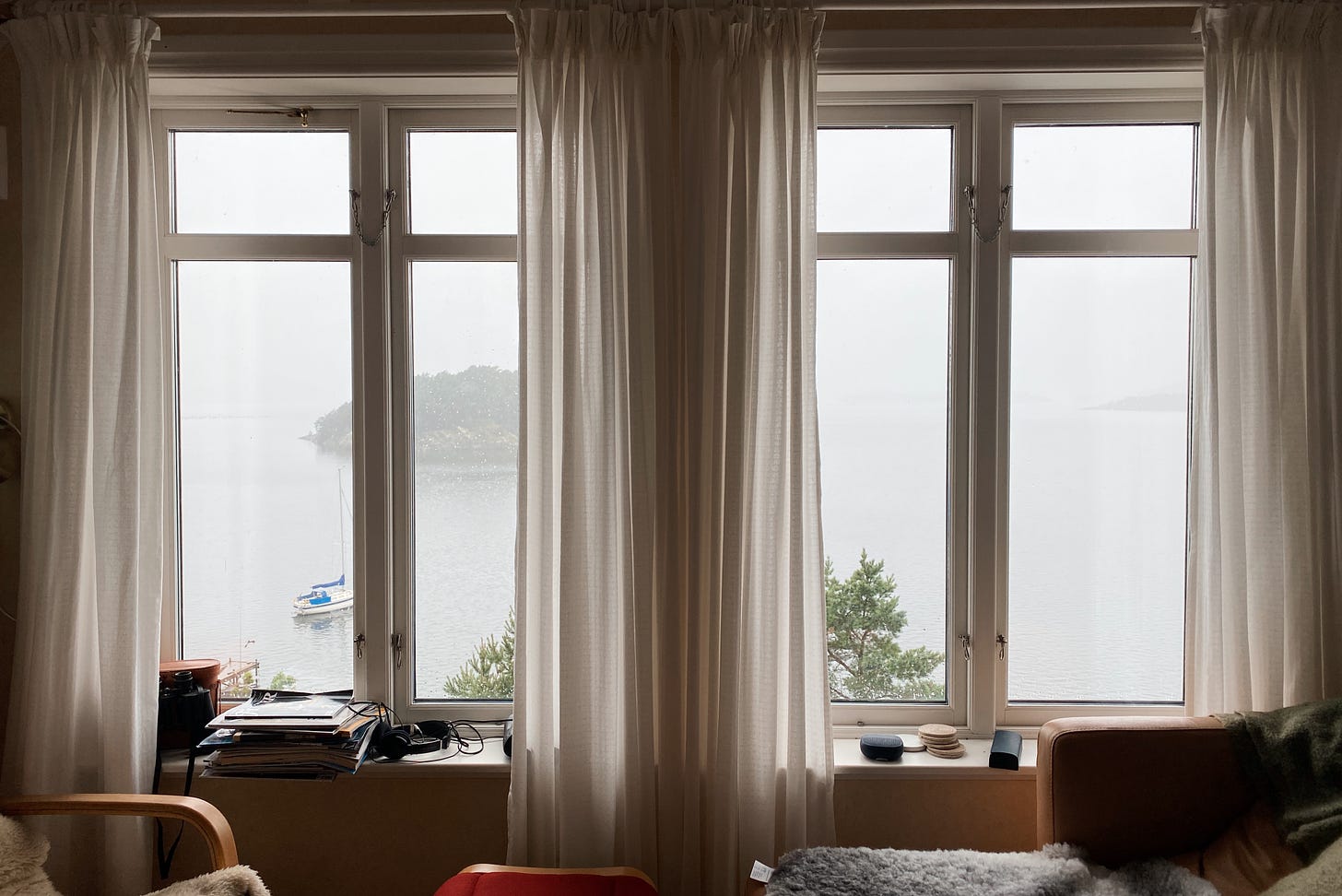 Imagem: Duas janelas altas com cortinas transparentes de onde se vê o mar, um barco e uma ilha