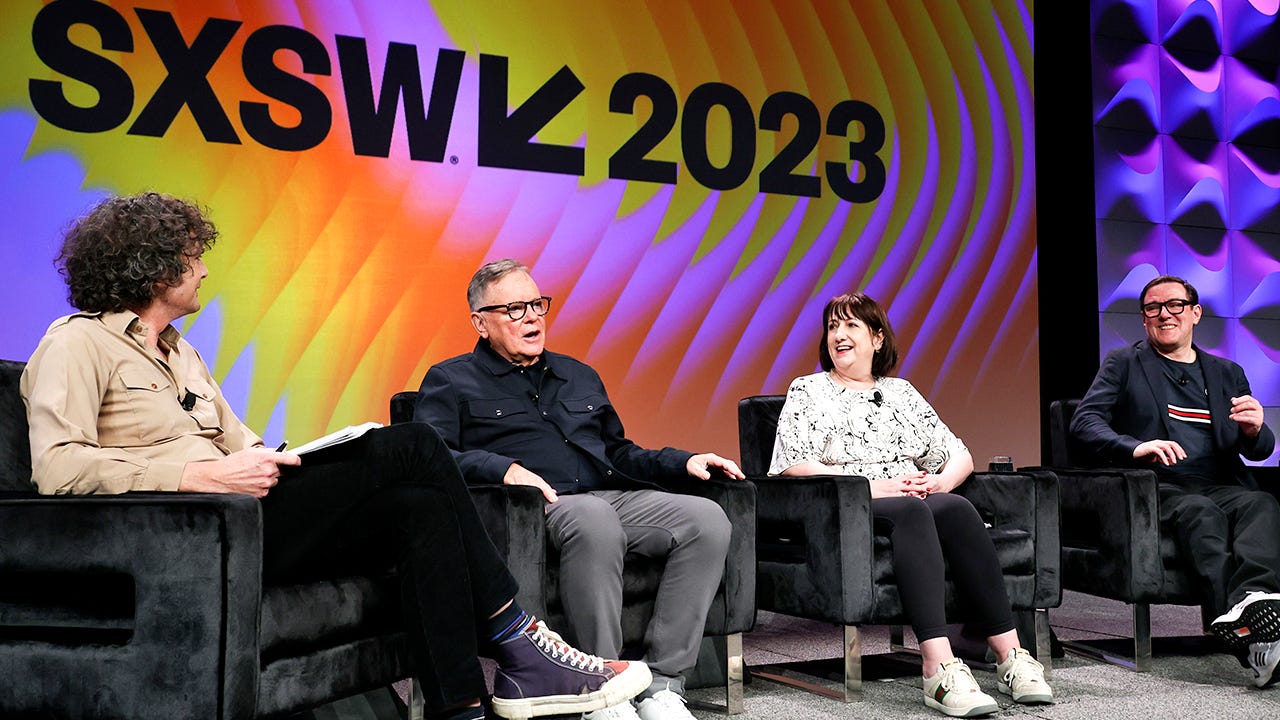 Jornalista entrevista a banda New Order. Estão todos sentados numa cadeira preta e atrás aparece uma imagem com SXSW 2023