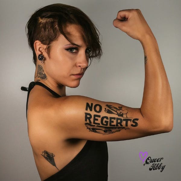 Woman with short brown hair and tattoos flexes her bicep with a tattoo that says "No Regerts" misspelled on her arm.