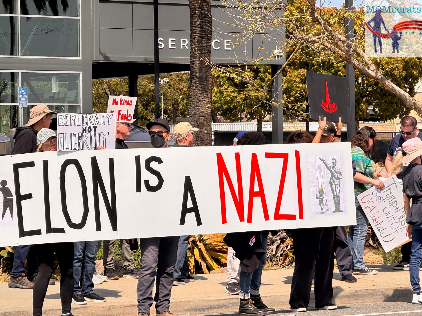 Protesters holding a long sign in front of the Camarillo Tesla dealership which says "Elon is a Nazi". Another protester holds a sign which is an upside down Tesla logo with two eyes cut out, making it look like a KKK hood.