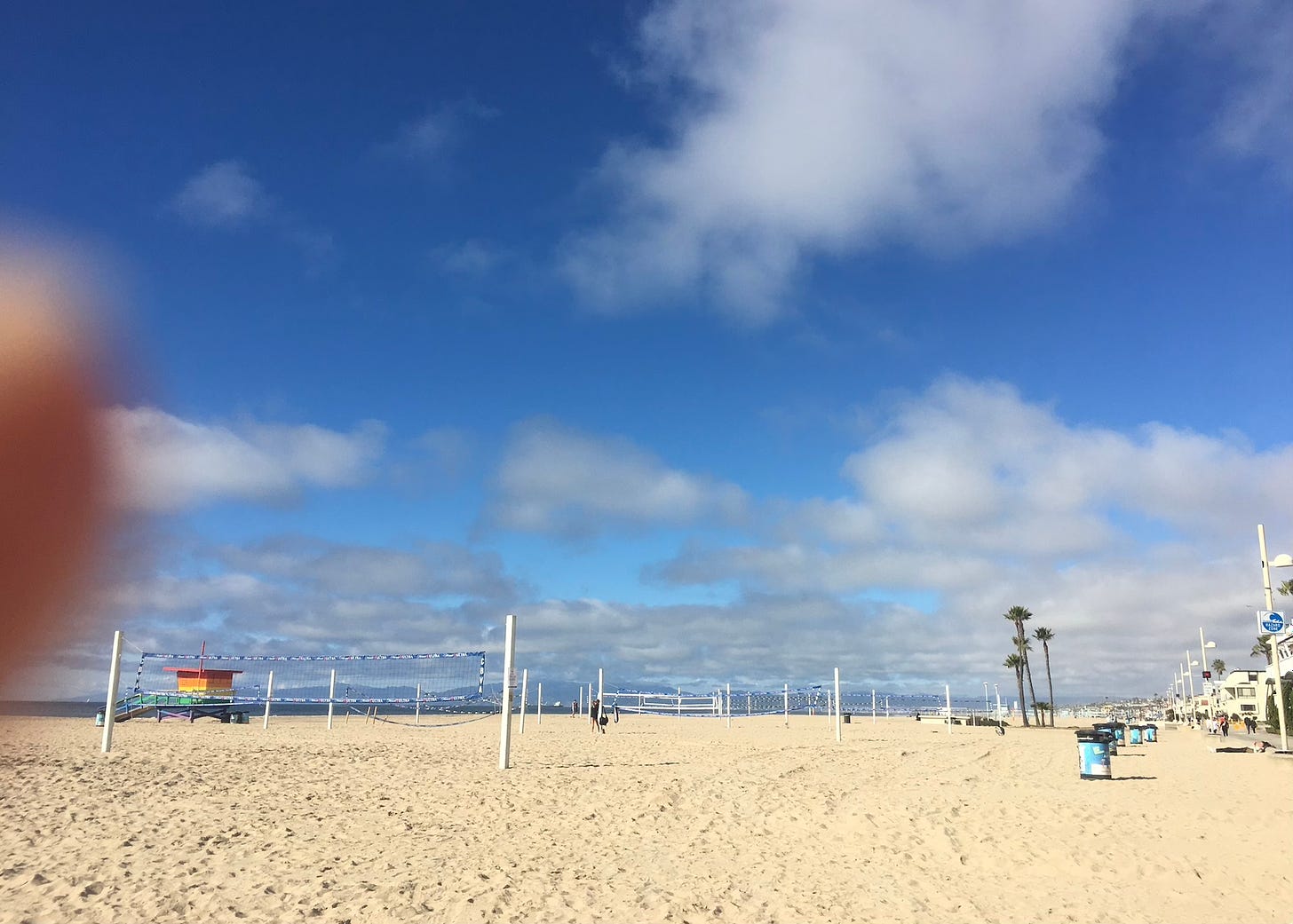 Very blue skies with a few clouds above a beach dotted with volleyball nets and palm trees. There is a blurry finger in the corner