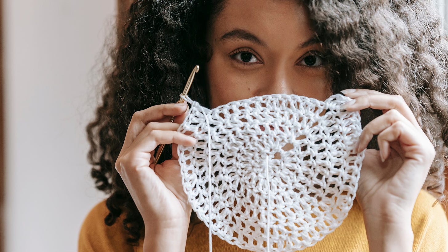 A woman with light brown skin and wavy black hair holds a partially completed white circle and crochet hook in front of her face. 