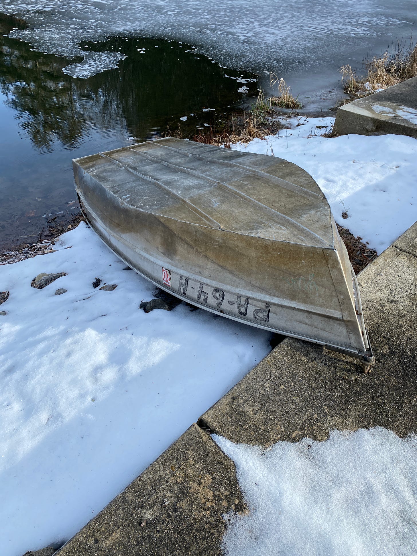 An overturned small rowboat on a snowy bank of a pond with skim ice