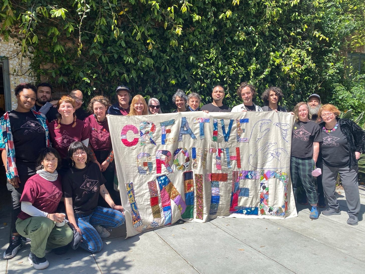 A photo of 18 people outside on a sunny day holding a large banner with quilted letters that say CREATIVE GROWTH UNITED.