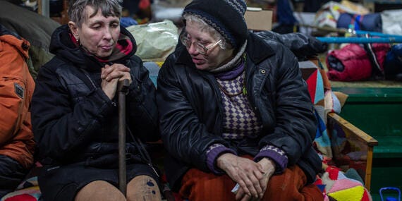 Two older women wearing winter clothes sit in a room.