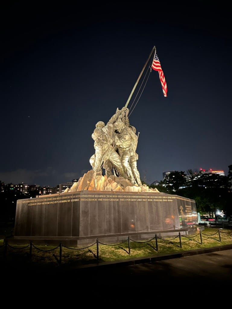 A statue of a person holding a flag with Marine Corps War Memorial in the background

Description automatically generated