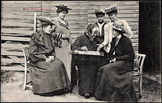 an old woman scrutinizes a spread of tarot cards while other women, well-dressed and younger, look on. The black and white photograph seems to be taken outside a barn, with the ladies sitting on wooden chairs at a little table. The picture was taken in Norway, perhaps in the late 19th century or early 20th.