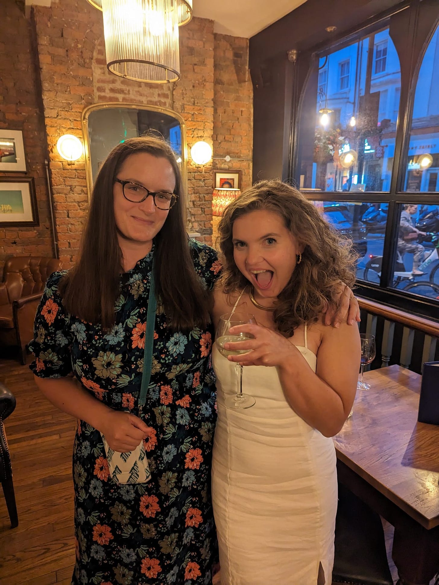 Mel and Fliss - Mel is a white woman with brown hair and glasses wearing a floral dress, Fliss is a white woman with wavy brown hair wearing a white dress and cheersing herself quite rightly