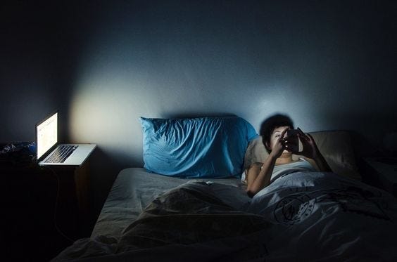 A human lays in bed on their phone. Beside them, their laptop brightens up a dark room.