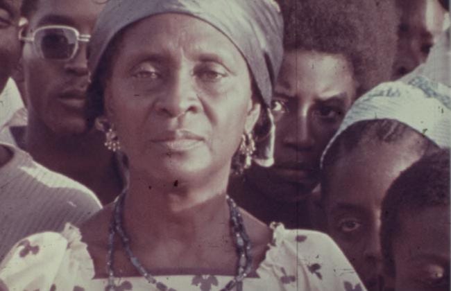 A group of people including children and adults stare at a scene not pictured in the frame. A woman in the center of the image is wearing gold earrings, blue beaded necklace and a silky headwrap.
