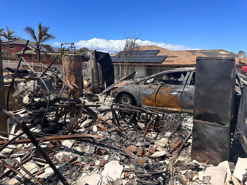 Color photo of a burned out home's garage strewn with wreckage, twisted metal. A car and water heater are easily recognizable, but as you look closer you can see a collaped shelving unit, weightlifting equipment, a bicycle frame, and canned food. In the background, some only lightly damaged homes are visible