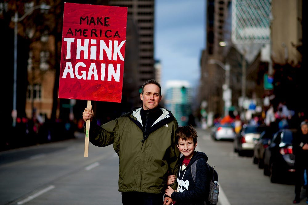 A man and a boy at a protest. The man is holding a sign that reads “Make America Think Again.”