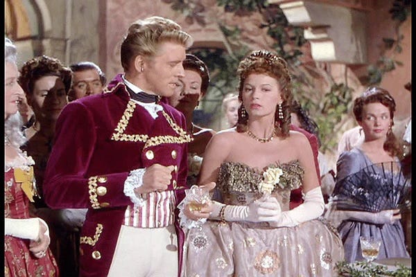 Burt Lancaster and Eva Bartok in The Crimson Pirate. He is impersonating Baron Gruda and she is wearing a ballgown. They are conversing while other party guests watch on from the background.