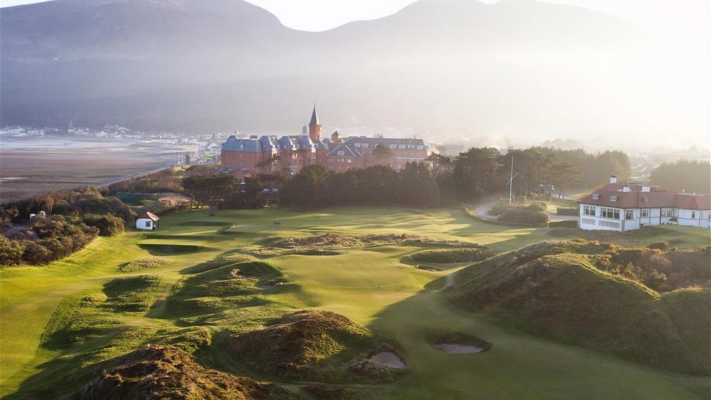 Morning view of Royal County Down Golf Course with dramatic sand dunes and undulating fairways in the foreground. A historic Irish town with prominent church spire sits nestled against misty mountains in the background. Golden morning light illuminates the links-style terrain while creating atmospheric shadows across the natural dunes. Morning view of Royal County Down Golf Course with dramatic sand dunes and undulating fairways in the foreground. A historic Irish town with prominent church spire sits nestled against misty mountains in the background. Golden morning light illuminates the links-style terrain while creating atmospheric shadows across the natural dunes.