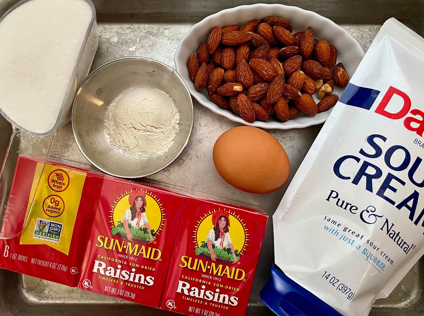 ingredients on a metal tray: boxes of raisins, sugar, flour, egg, sour cream, and nuts