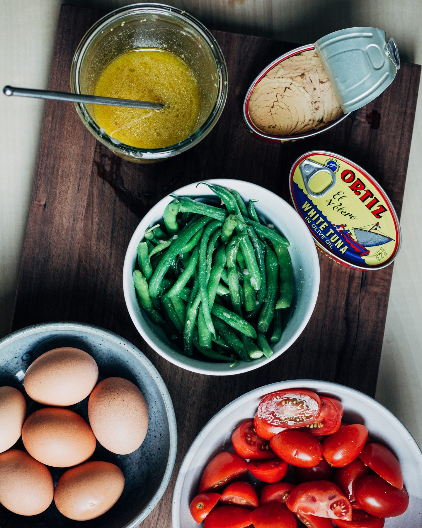 A cutting board with a jar of vinaigrette, bowls of green beans, tomatoes, and eggs, and canned tuna. 