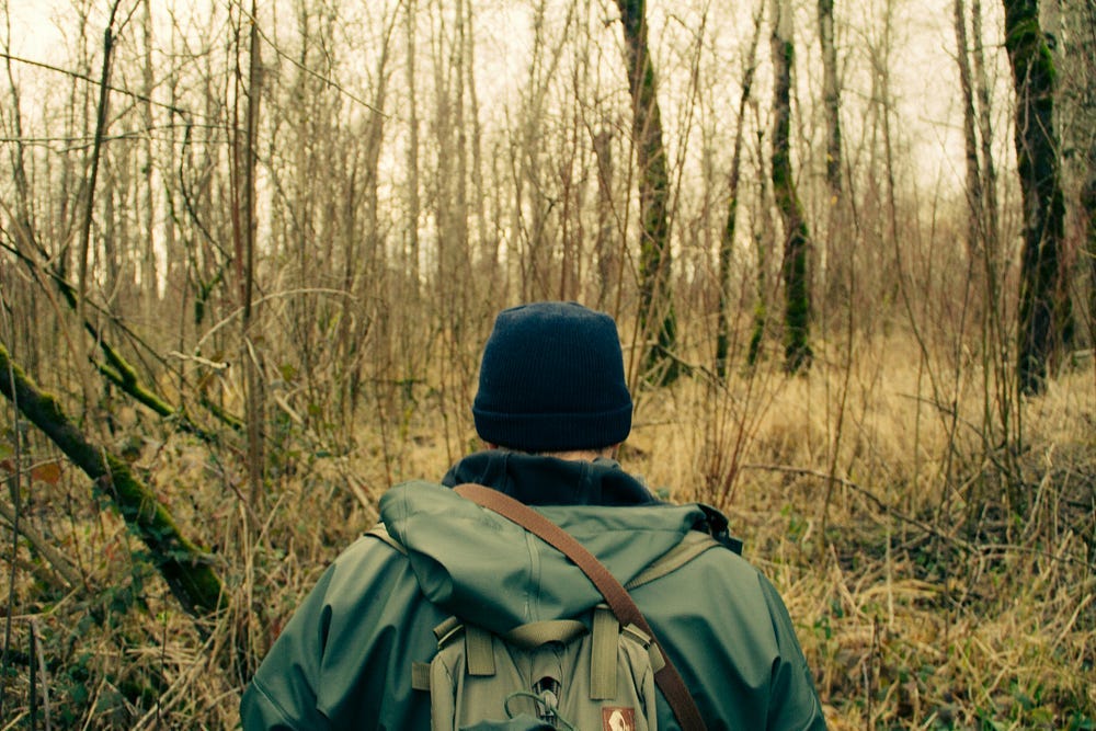 A man in the woods, wearing a black cap, backpack and a green jacket.