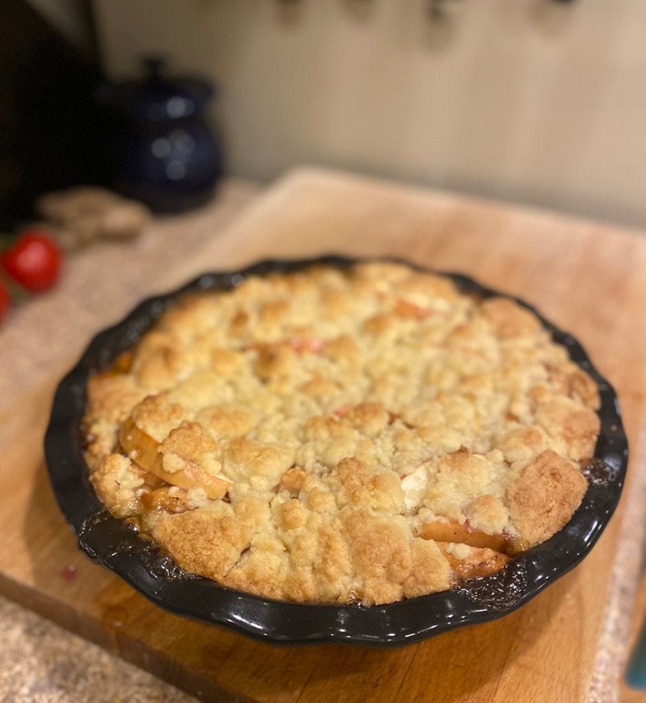 A black pie dish full of apple cobbler, the top browned and crumbly, sitting on a wood cutting board.