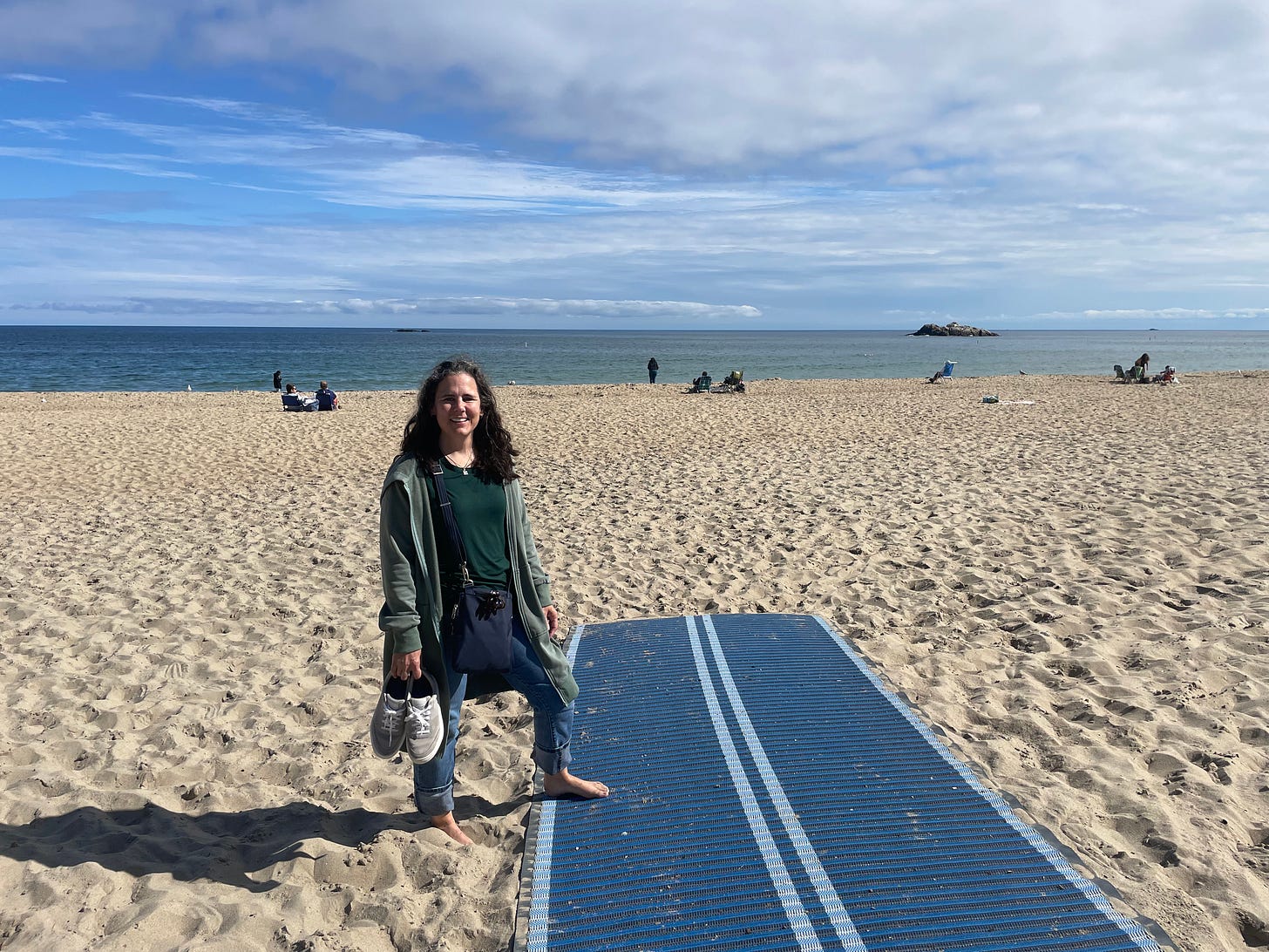 In the background is a flat horizon; a blue sky with white thin clouds over still, teal-blue water. A few people are dotting the flat, beige sand beach in the background.In the foreground: The author standing in jeans, green t-shirt, and a long green sweater. She's holding shoes in one hand; she is barefoot and one foot is resting on a dark blue mat with a light blue double line down the middle. 