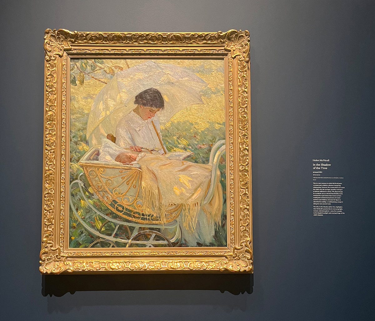 Framed painting of a mother and baby sitting outside in the shade. The mother holds a white parasol with her hand on the baby's pram but her focus on the book she's reading. The baby appears to be asleep in the ornately decorated pram and there's a light golden coloured blanket with long trailing fringe draped over the baby.