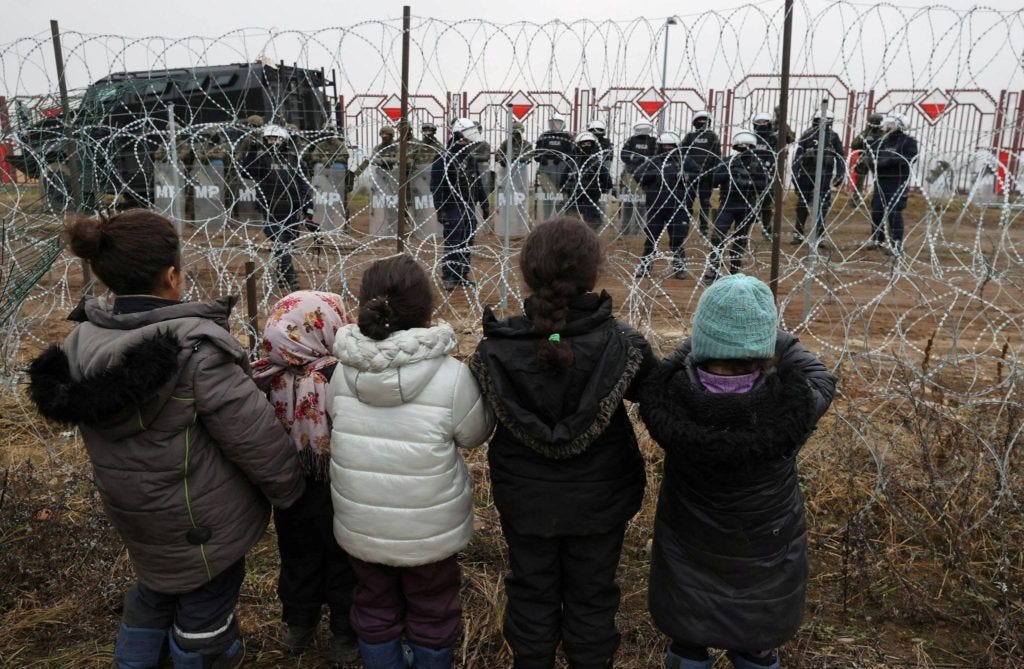 Children look through barbed wire fence at border guards with shields and armour